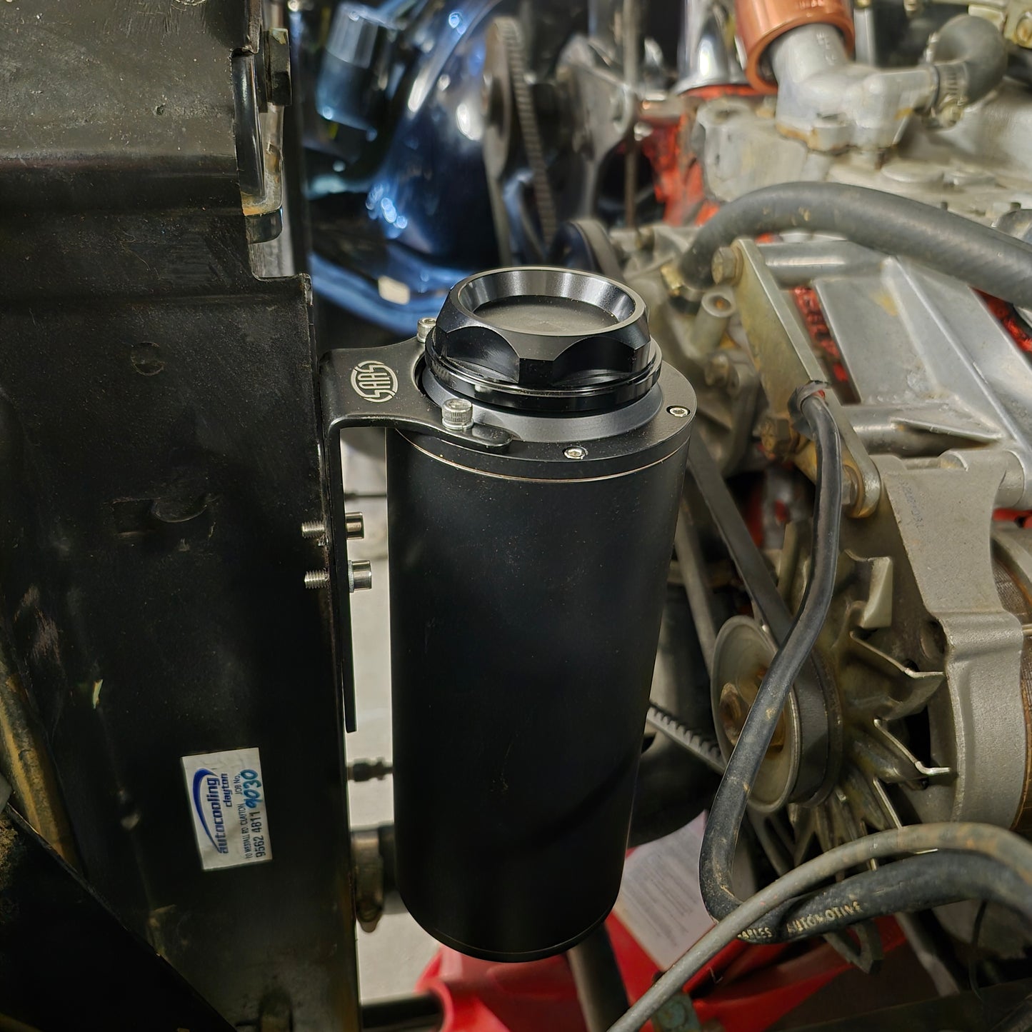 Close-up of a SAAS Water Overflow Tank on a vehicle engine with visible branding.
