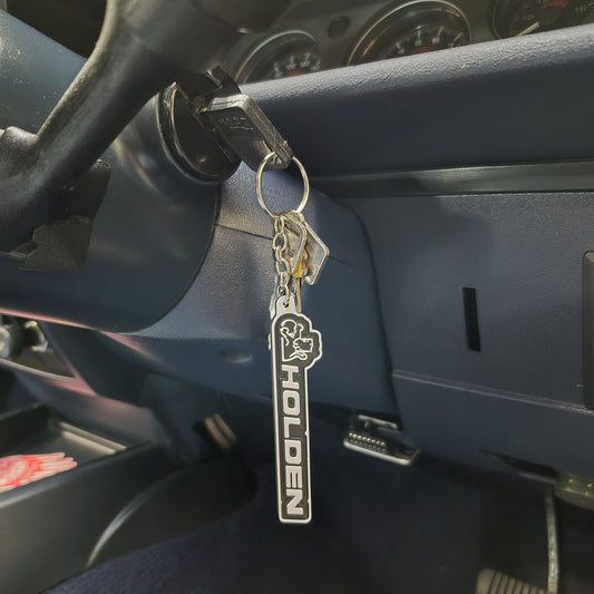 A silver metal car keychain with a black and white vertical 'HOLDEN' logo and a horse emblem, hanging from a key in a vehicle's ignition.