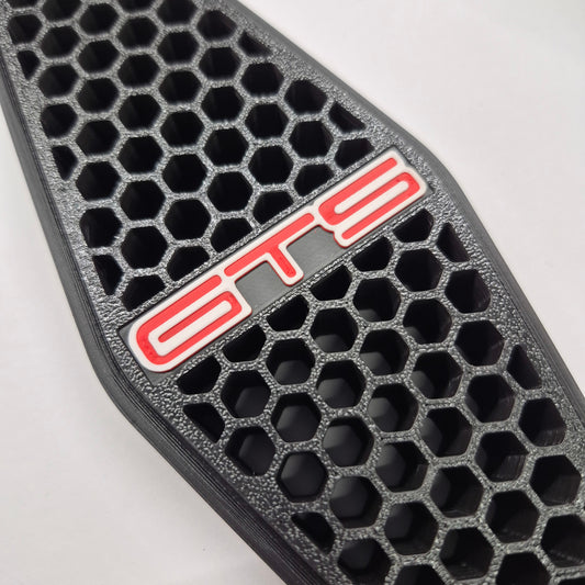 A close-up of a black honeycomb-patterned grille insert with a red and white 'GTS' badge on a light gray background.
