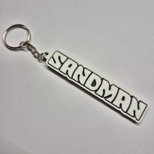 A white and black rubber keychain featuring the word 'SANDMAN' in a bold, stylized comic-style font, attached to a silver metal split ring and chain on a plain white background.