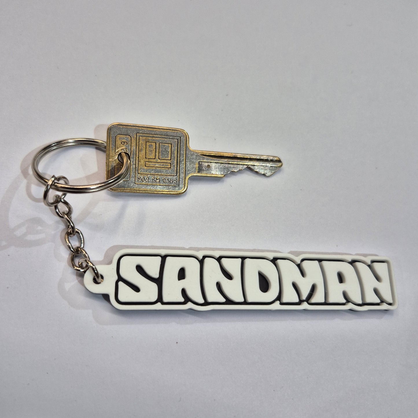 A novelty keychain featuring a silver metal key and chain attached to a white rubber tag with the word 'SANDMAN' in bold black letters.