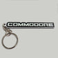 A black and silver metallic keychain featuring the word 'COMMODORE' in bold white letters on a black background, attached to a silver split key ring.