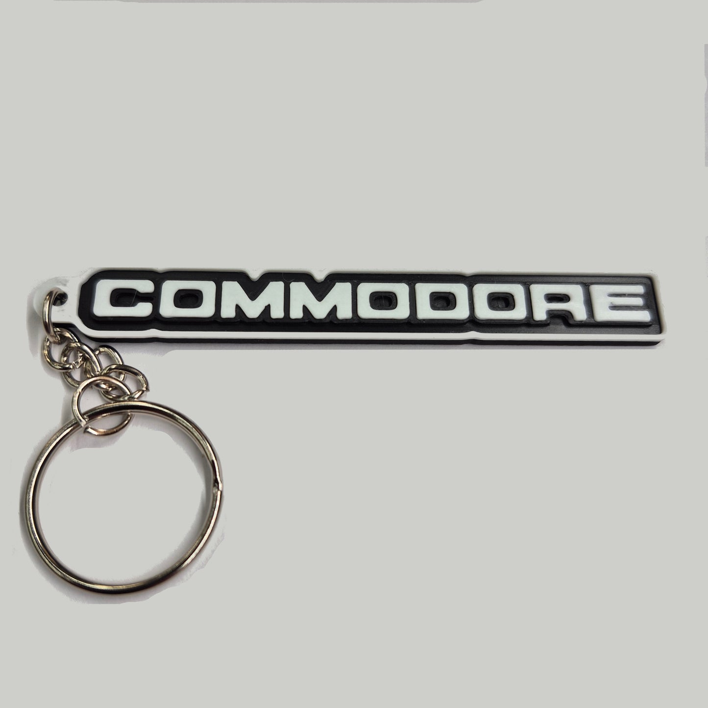 A black and silver metallic keychain featuring the word 'COMMODORE' in bold white letters on a black background, attached to a silver split key ring.
