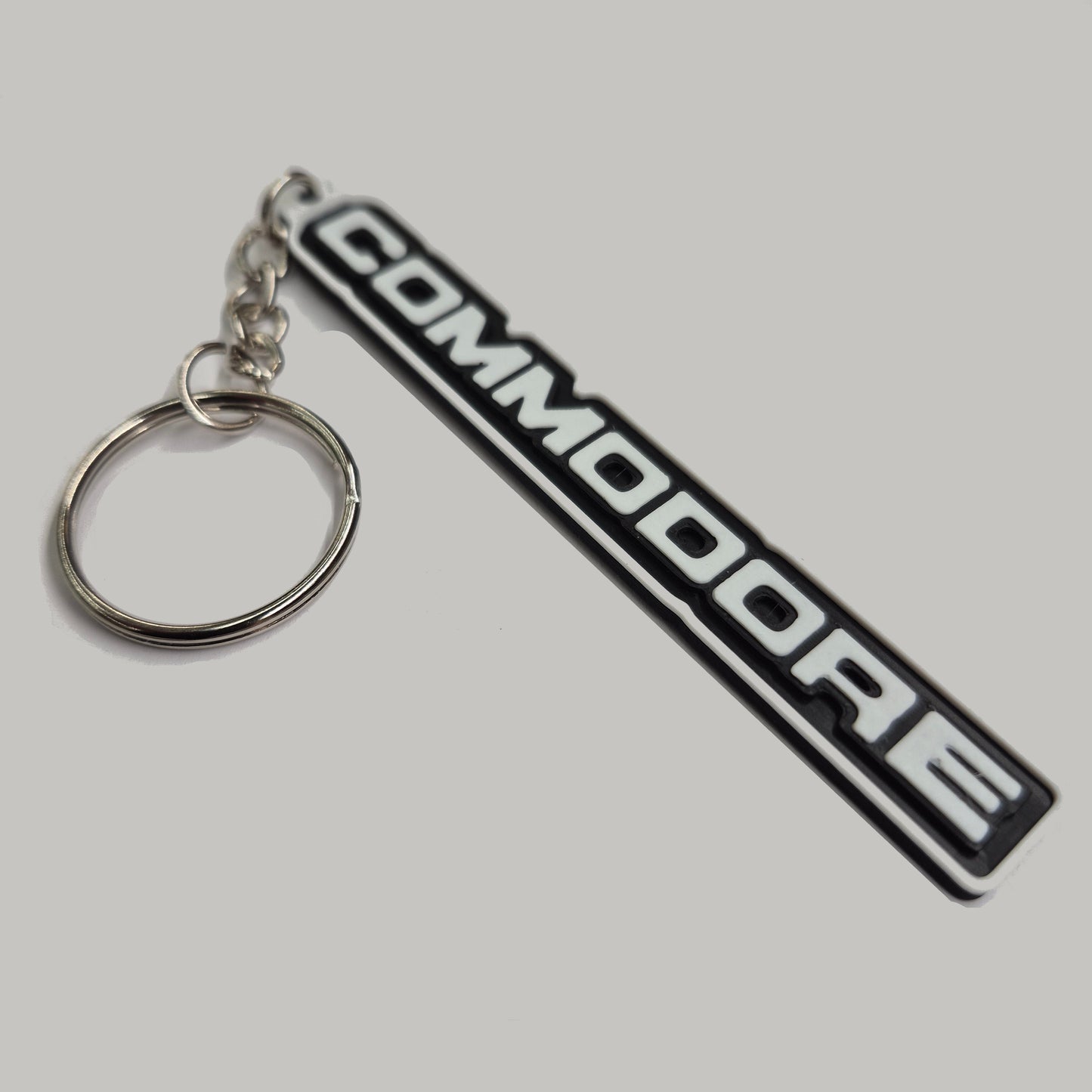 A black and white rubber keychain featuring the 'COMMODORE' logo from a classic car, attached to a silver metal split ring and chain, set against a plain light gray background.