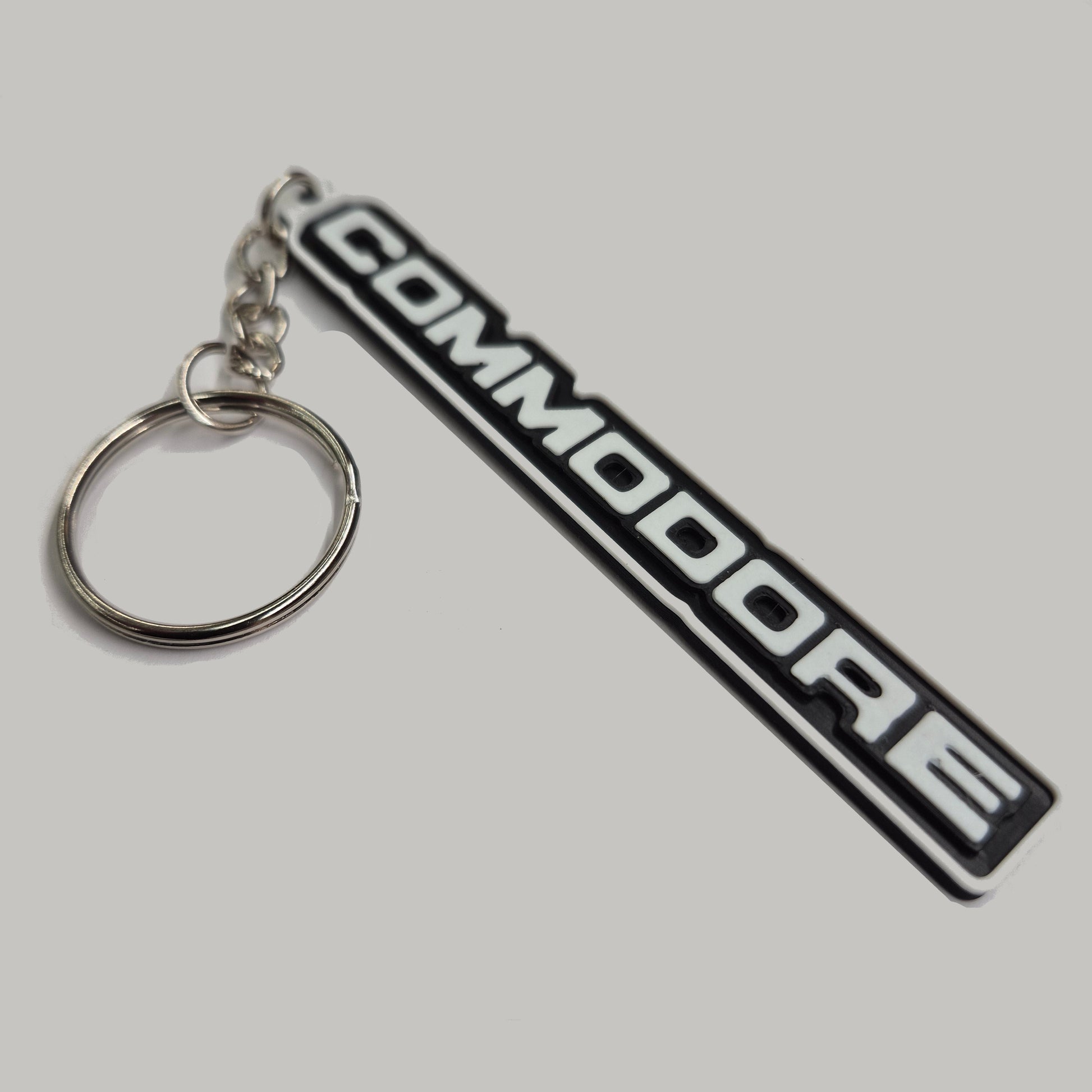 A black and white rubber keychain featuring the 'COMMODORE' logo from a classic car, attached to a silver metal split ring and chain, set against a plain light gray background.