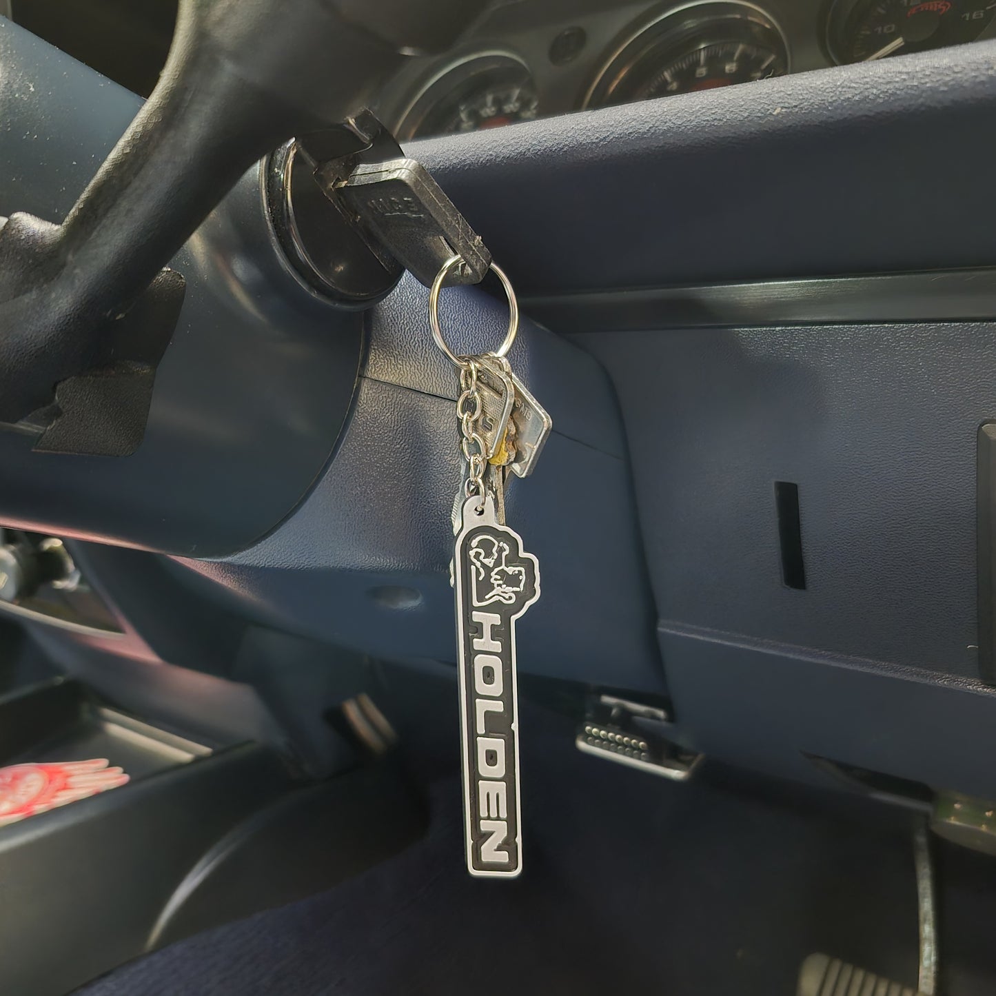 A silver metal car keychain with a black and white vertical 'HOLDEN' logo and a horse emblem, hanging from a key in a vehicle's ignition.