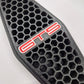 A close-up of a black honeycomb-patterned grille insert with a red and white 'GTS' badge on a light gray background.