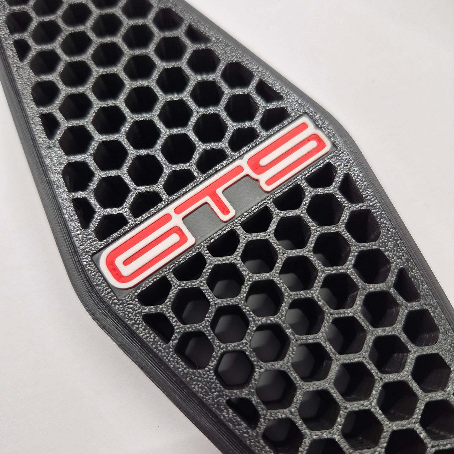 A close-up of a black honeycomb-patterned grille insert with a red and white 'GTS' badge on a light gray background.
