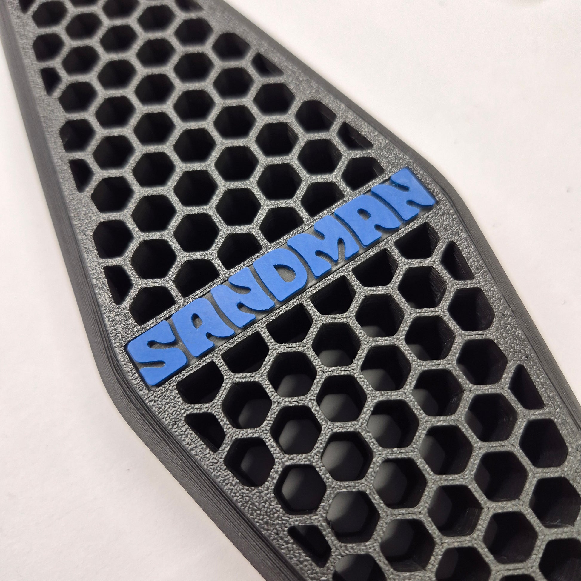 A close-up of a black honeycomb-patterned textured pad with a blue raised 'SANDMAN' logo, shown on a white background.