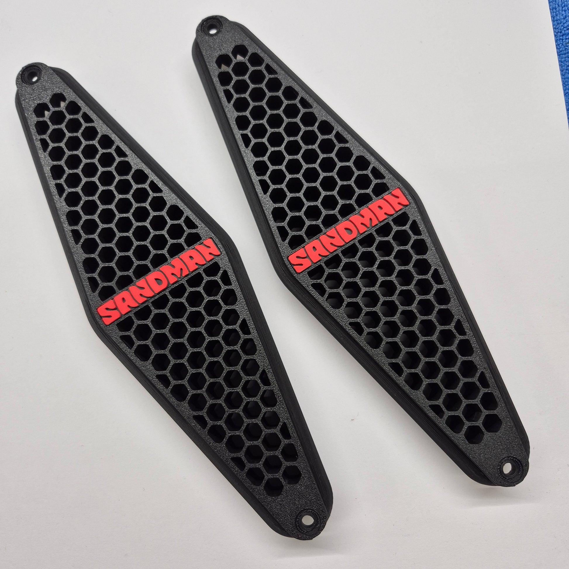 A pair of black motorcycle tank covers with a honeycomb pattern and red 'SANDMAN' branding, displayed on a white background.