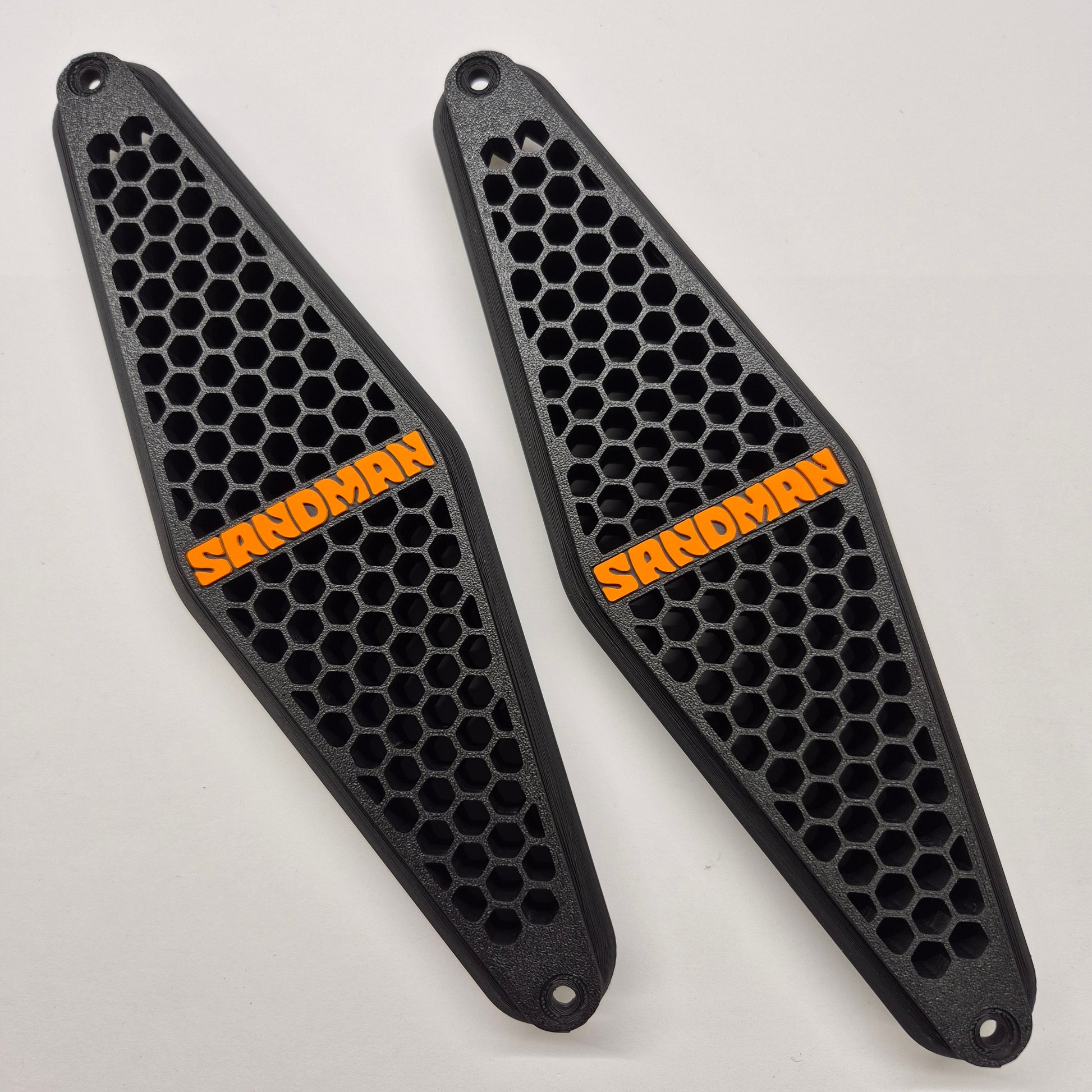 Two black honeycomb-patterned sandboard/skating pads with orange 'SANDMAN' branding, displayed on a light gray background.