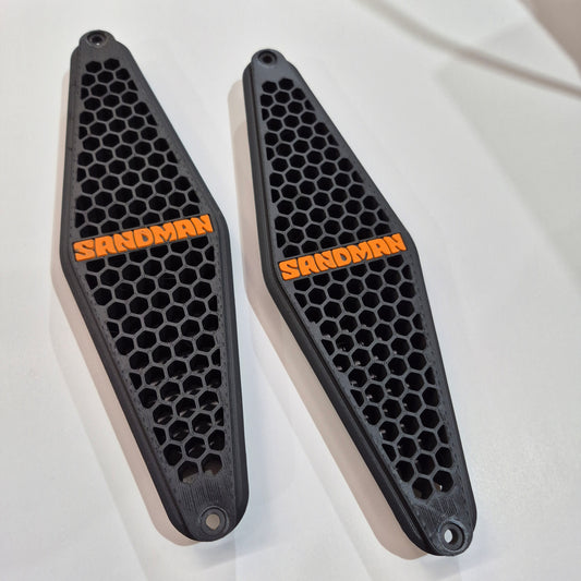 Two black honeycomb-patterned motorcycle foot pegs with orange branding on a white background