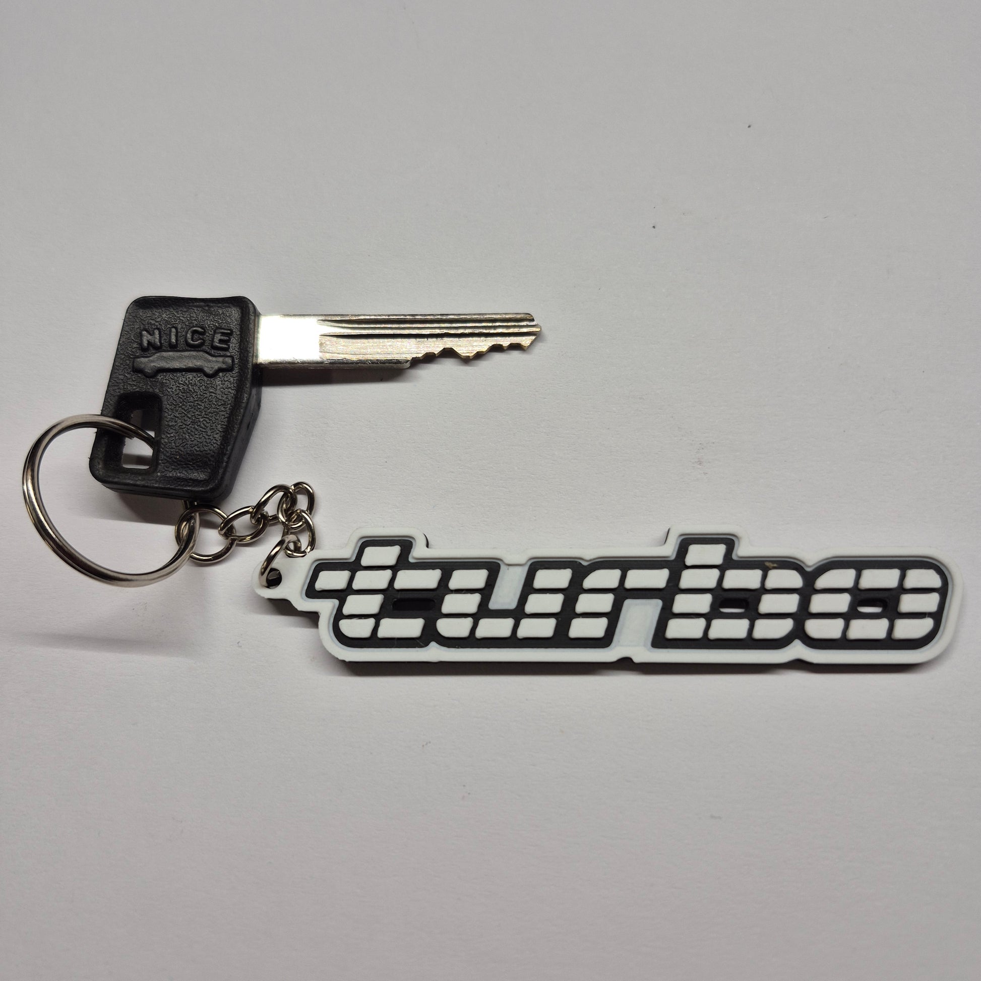 A black and white keychain featuring a stylized 'MUGUE' logo, shown with a metal key and keyring on a light gray background.