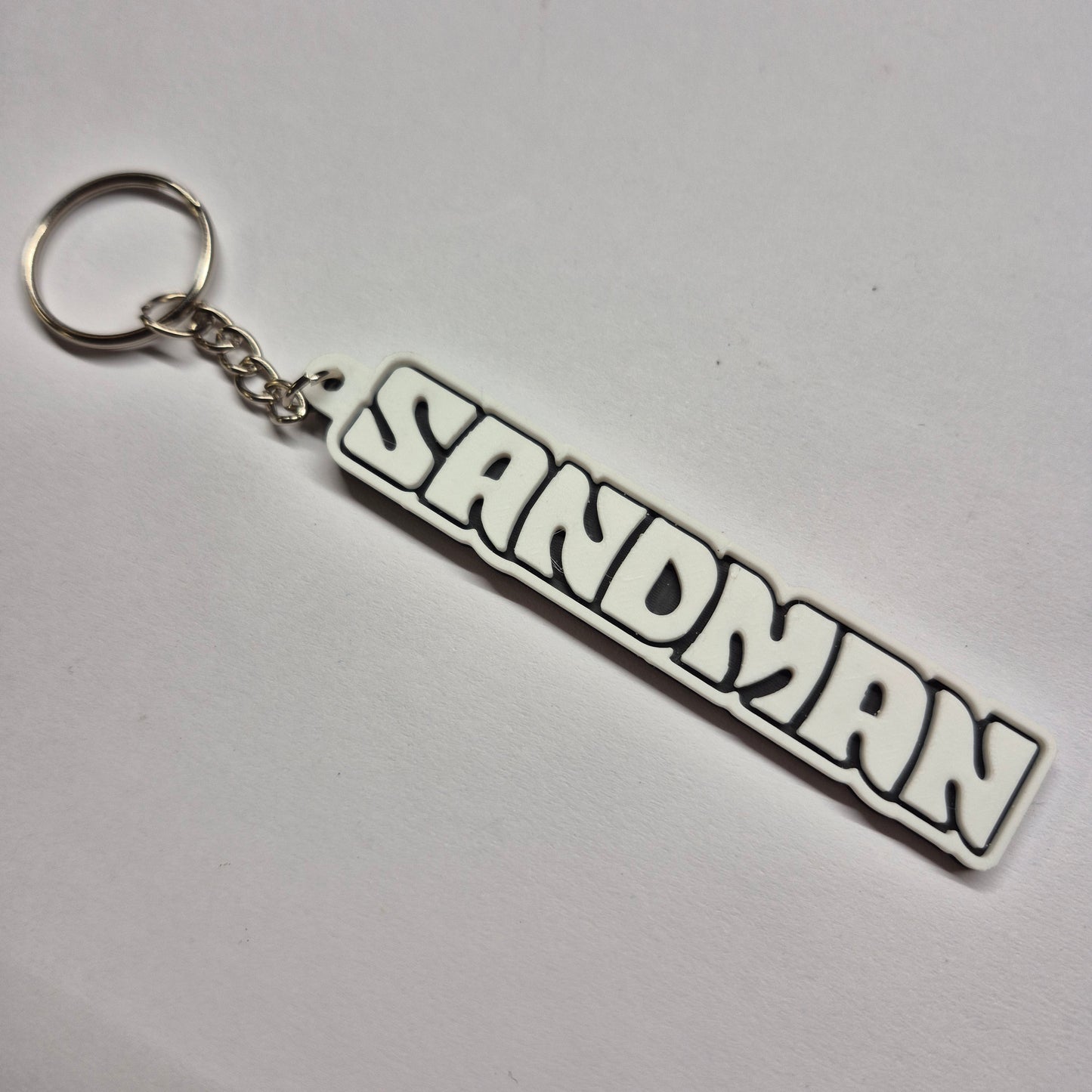 A white and black rubber keychain featuring the word 'SANDMAN' in a bold, stylized comic-style font, attached to a silver metal split ring and chain on a plain white background.
