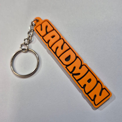 An orange and black rubber keychain featuring the word 'SANDMAN' in a bold, stylized font, attached to a silver metal split ring on a light gray background.