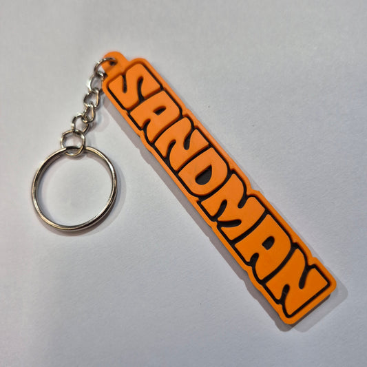 An orange and black rubber keychain featuring the word 'SANDMAN' in a bold, stylized font, attached to a silver metal split ring on a light gray background.