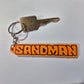 A vintage-style metal key with a round keyring attached next to an orange rectangular keychain featuring the word 'SANDMAN' in bold black letters, shown on a plain light background.