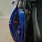Close-up of a blue motorcycle fender with a black vented grille featuring the red 'SHEDMAN' logo, next to a black leather seat.