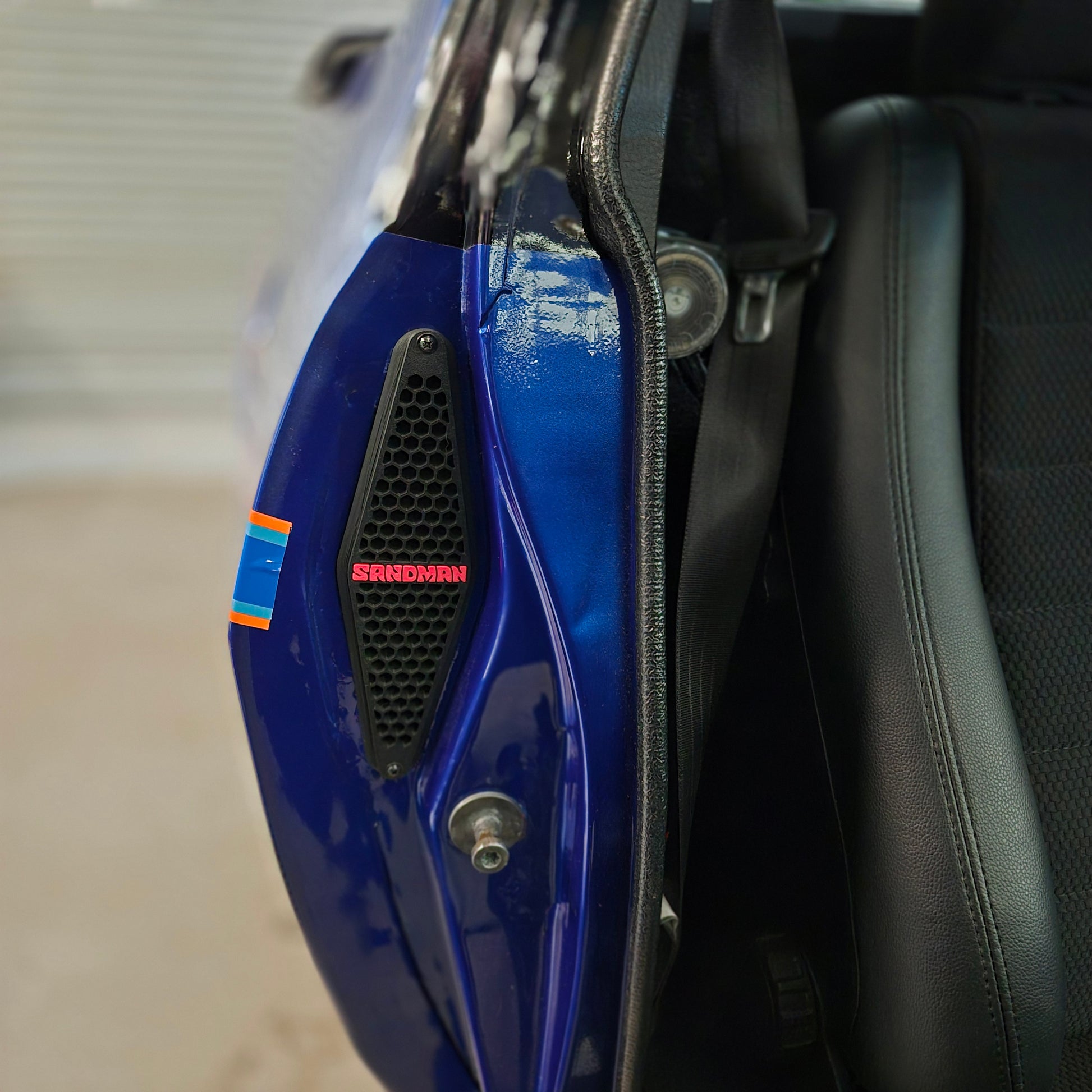 Close-up of a blue motorcycle fender with a black vented grille featuring the red 'SHEDMAN' logo, next to a black leather seat.