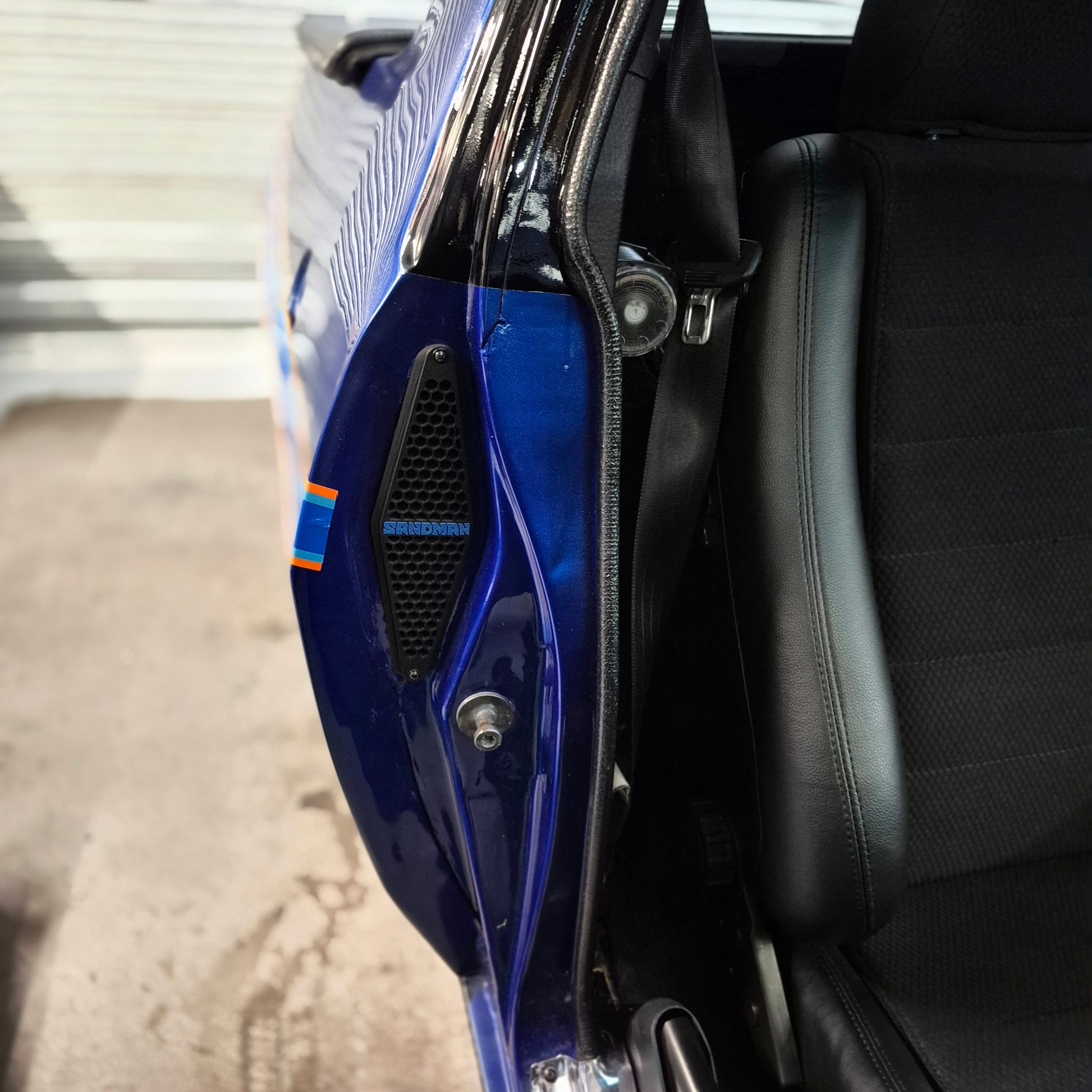 Close-up of a blue car door panel with a black speaker grille and a small orange accent stripe, next to a black leather seat.
