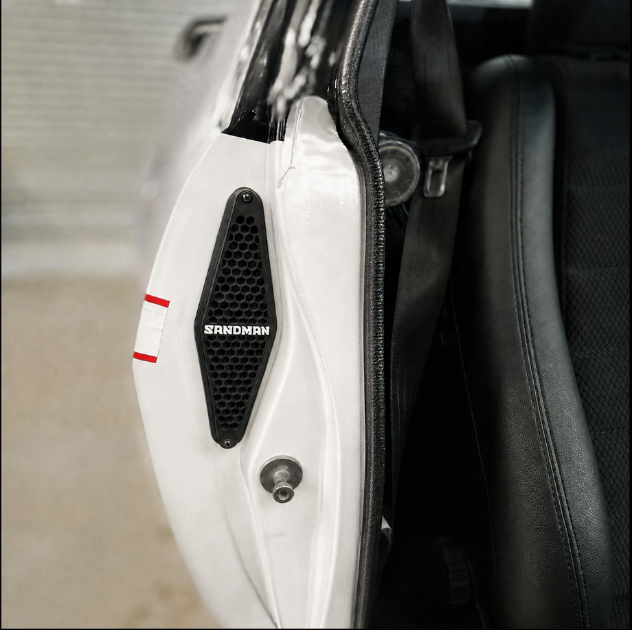 Close-up of a white vehicle door panel with a black mesh speaker grille labeled SANDMAN, next to a black leather seat.