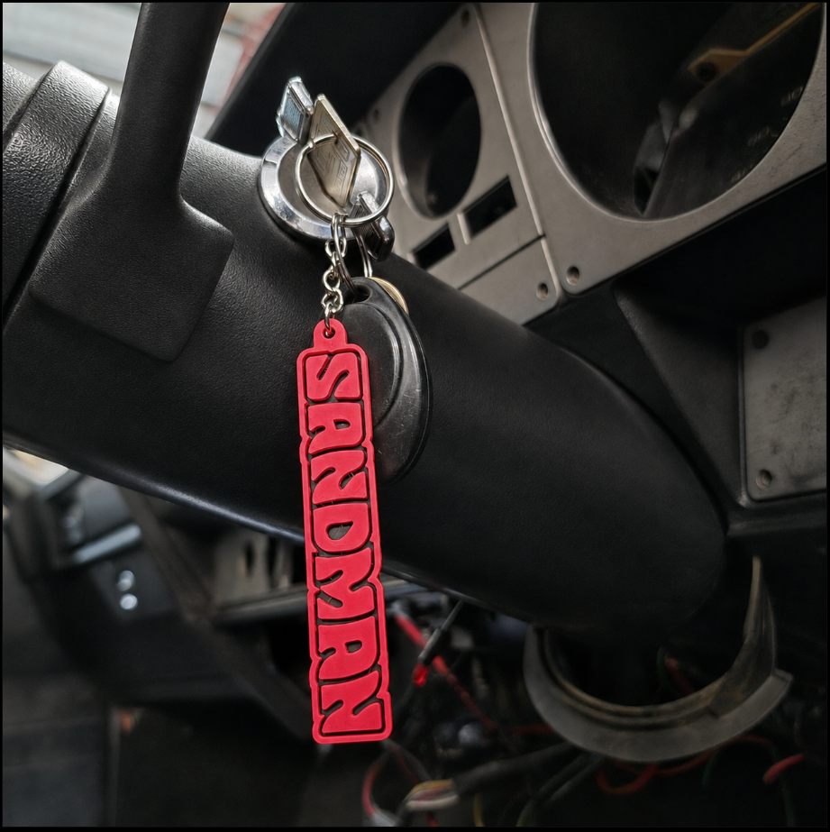 A red and black vertical keychain featuring the word 'SANDMAN' in raised letters, hanging from a key in a car ignition.