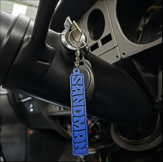 A blue and black vertical keychain featuring the word 'SANDMAN' in raised letters, hanging from a key in a car ignition.