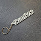 A silver metal keychain featuring a stylized 'turbo' wordmark with a checkered flag motif, attached to a split key ring and resting on a dark textured surface.