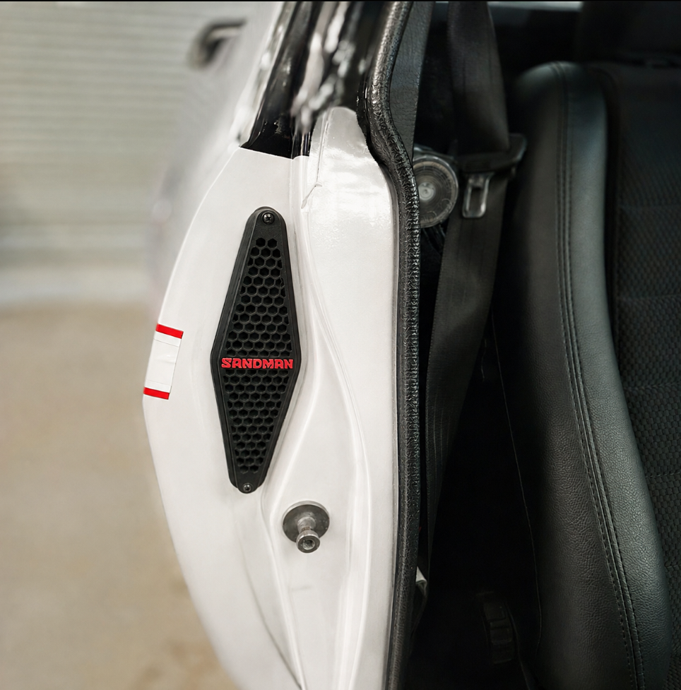 Close-up of a white vehicle door panel with a black mesh grille featuring the red SANDMAN logo, next to a black leather seat.
